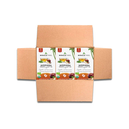 Open cardboard box containing three packages of Miracle Tree Organic Moringa Tea, Apple & Cinnamon. The packaging features images of apples, cinnamon, and tea leaves with a superfood theme in green and white design elements.