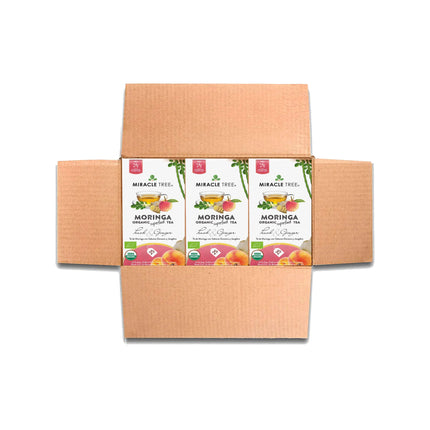 The open cardboard box holds four boxes of Miracle Trees Organic Moringa Tea, Peach & Ginger. This caffeine-free superfood features vibrant peach and ginger illustrations on each box front.