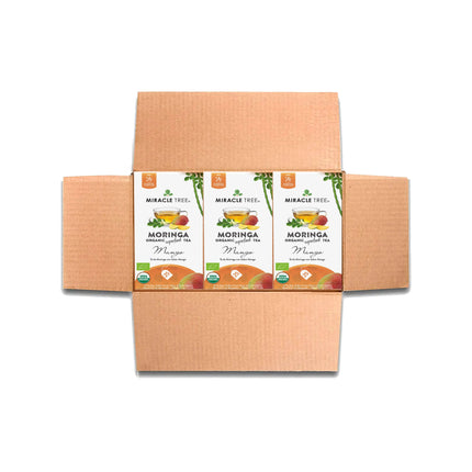 Open cardboard box with three packs of Miracle Trees Organic Moringa Tea in Mango flavor. Each pack displays a cup of tea with fruit graphics, highlighting this superfood, and features green and orange accents.