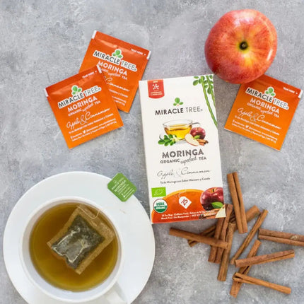 A flat lay of Miracle Tree Organic Moringa Tea, Apple & Cinnamon features a superfood-rich tea bag in a cup surrounded by its tea packets, an apple, the signature box, and cinnamon sticks on a gray surface.