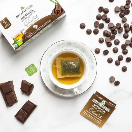 A top-down view shows a caffeine-free cup made from a Miracle Tree Organic Moringa Tea bag in Chocolate flavor, surrounded by its box and packet, chocolate pieces, and scattered chips on a white background.