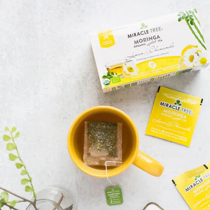 A yellow mug with a tea bag sits beside a Miracle Tree box and packet of Organic Moringa Tea, Lemon & Chamomile, a caffeine-free superfood blend. A small leafy branch adds decoration.