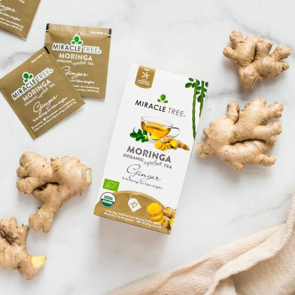 A box of Miracle Tree’s Organic Moringa Tea, Ginger flavor, rests on a marble surface surrounded by fresh ginger roots and two tea packets, with a beige cloth visible at the bottom corner.