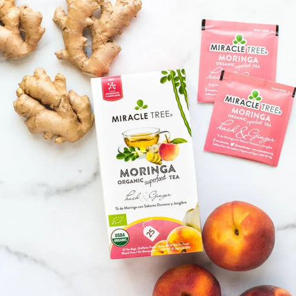 A box of Miracle Trees Organic Moringa Tea, Peach & Ginger, caffeine-free and packed with superfood benefits, is elegantly arranged with two sachets, fresh ginger roots, and three juicy peaches on a pristine white surface.