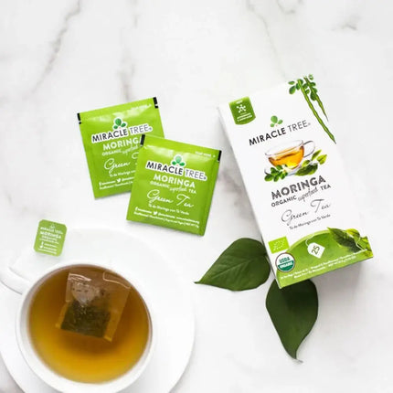 On a white surface, a box with two packets of Miracle Tree Organic Moringa Tea, Green Tea sits beside a brewed cup with a dangling tea bag tag. Two green leaves are next to the cup.