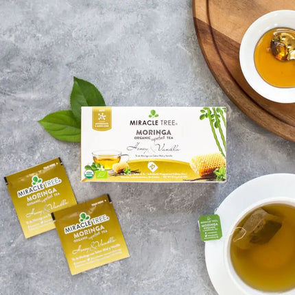 A box of Miracle Trees Organic Moringa Tea, Honey & Vanilla sits on the table. Nearby, two caffeine-free tea packets and a steaming cup await indulgence. A wooden board with honey and green leaves completes this vibrant superfood scene.