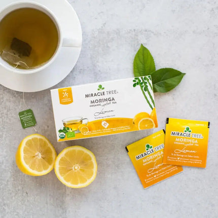 A cup of caffeine-free tea sits beside a box of Miracle Tree Organic Moringa Tea, Lemon flavor, two lemons, and two lemon-flavored tea packets. Fresh green leaves are artfully displayed on a light gray surface.