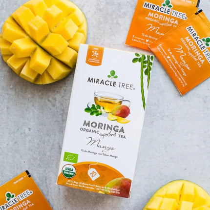 A Miracle Tree Organic Moringa Tea, Mango box is surrounded by mango slices and three sachets on a gray surface. This organic-certified, caffeine-free superfood blend displays mango and moringa leaf images.