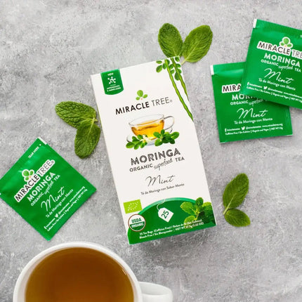 A box of Miracle Tree Organic Moringa Tea, Mint is surrounded by three green tea packets and fresh mint leaves, with a teacup of brewed caffeine-free tea in the lower left corner on a gray surface.
