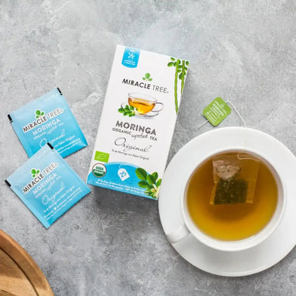 A box of Miracle Tree Organic Moringa Tea, Original with two sachets is next to a white cup of the brew, with the tea tag resting on the saucer against a textured gray background.