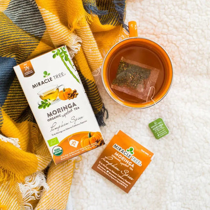 A box of Miracle Trees Organic Moringa Tea, Pumpkin Spice, rests beside a steaming caffeine-free cup with the bag inside. Nearby, a tea packet and a green-yellow textured blanket on a light surface enhance the cozy charm of this superfood experience.