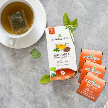 A cup of nutrient-rich Organic Moringa Tea, Rooibos by Miracle Tree, sits on a grey surface with a teabag on a saucer. Nearby, the tea box is surrounded by three sachets and decorated with scattered green leaves.