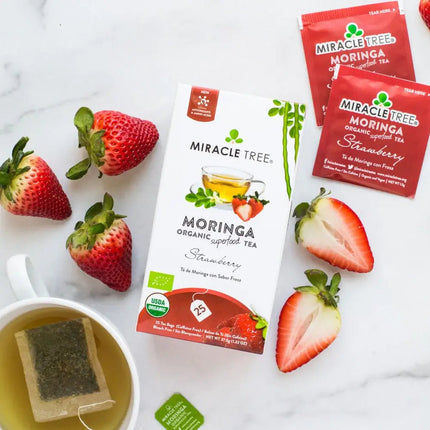 A box of Miracle Tree Organic Moringa Tea, Strawberry flavor, sits with fresh strawberries and tea bags. A brewed cup with a tea bag is visible. The vibrant packaging showcases strawberries and green accents, highlighting this delightful caffeine-free superfood tea.
