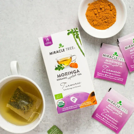 A box of Miracle Trees Organic Moringa Tea, Turmeric flavor, is surrounded by tea bags and turmeric powder beside a white mug of the superfood infusion on a light gray surface.