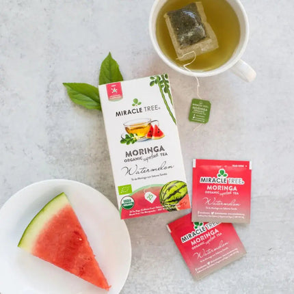 A slice of watermelon, a Miracle Tree Organic Moringa tea packet, a caffeine-free teacup with a teabag, and two tea packets are arranged on a light surface with a decorative green leaf.